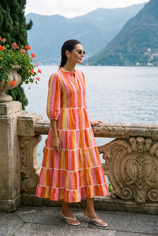 A woman wearing a tiered midi dress with vertical pink, orange, and yellow stripes stands on a stone terrace overlooking a scenic lake and mountains.