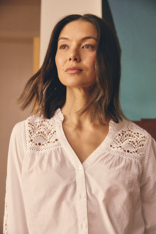 Woman in front of an artwork, indoors wearing white shirt with lace details at the shoulder. 