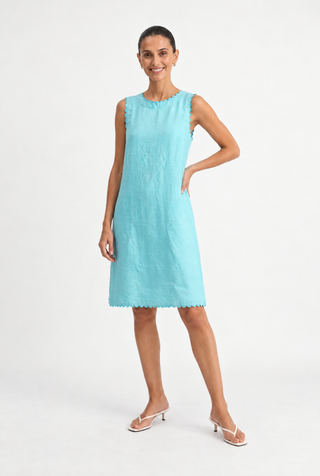 A woman modeling a sleeveless light blue knee-length dress with a scalloped hem and subtle textured pattern, paired with white heeled sandals, against a plain white studio background.